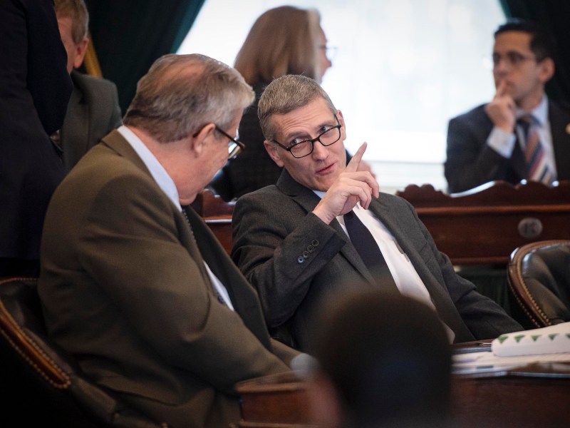 Two men in suits are seated, having a discussion. One gestures with his finger while speaking. Other people are visible in the background.