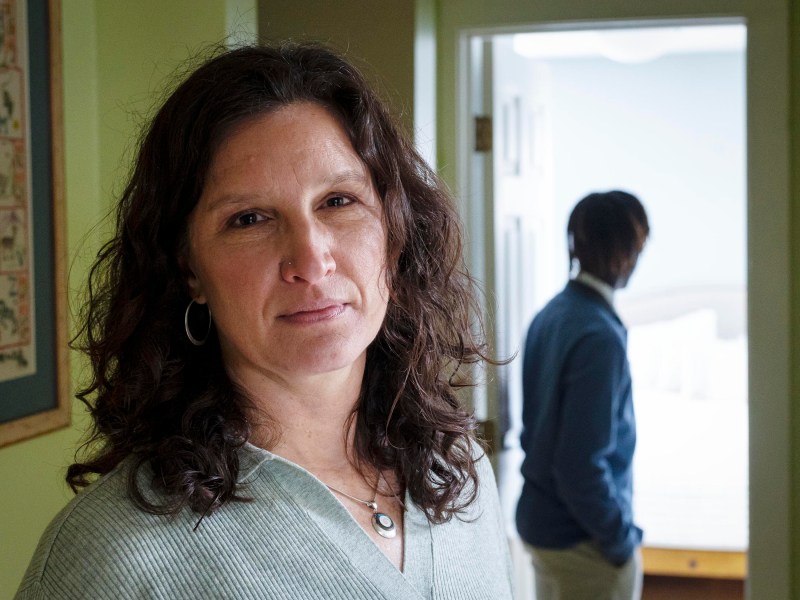 Woman with curly hair stands in focus while another person in a blue sweater is blurry in the background, standing near a door.