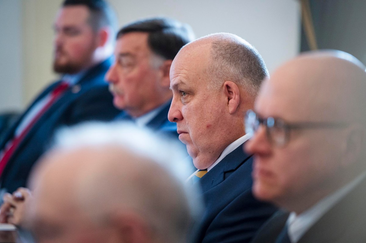 A row of men in formal attire sit attentively, facing left. The foreground man is bald and wearing glasses.