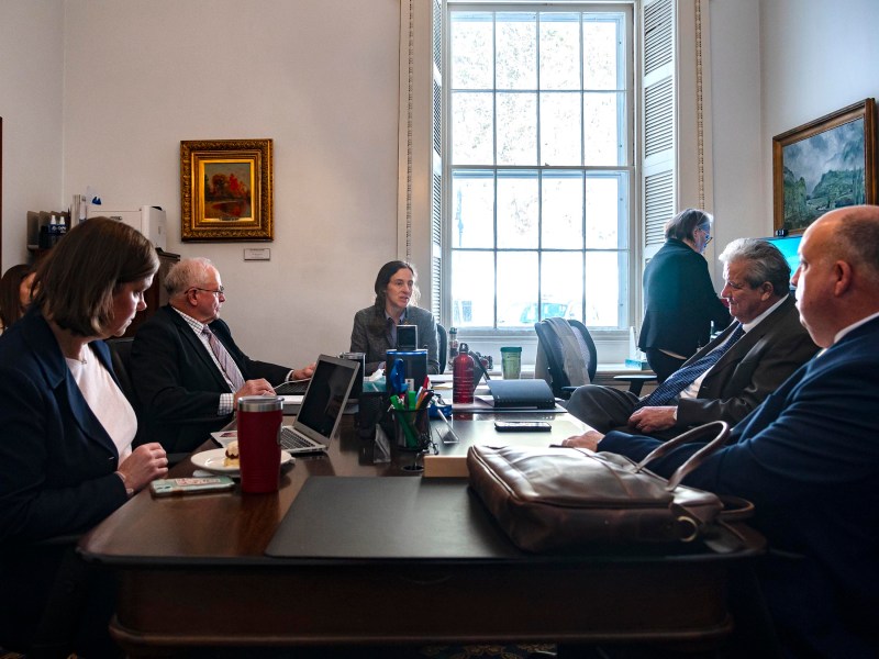 A group of people in business attire sit around a conference table with papers, laptops, and beverages in an office with large windows and framed paintings.