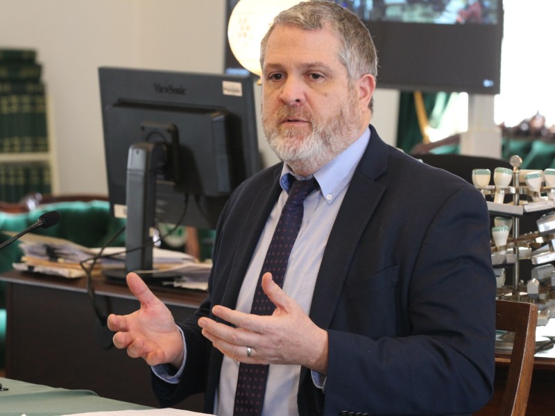 A man with a beard speaks while seated at a desk with a computer monitor and books in the background.