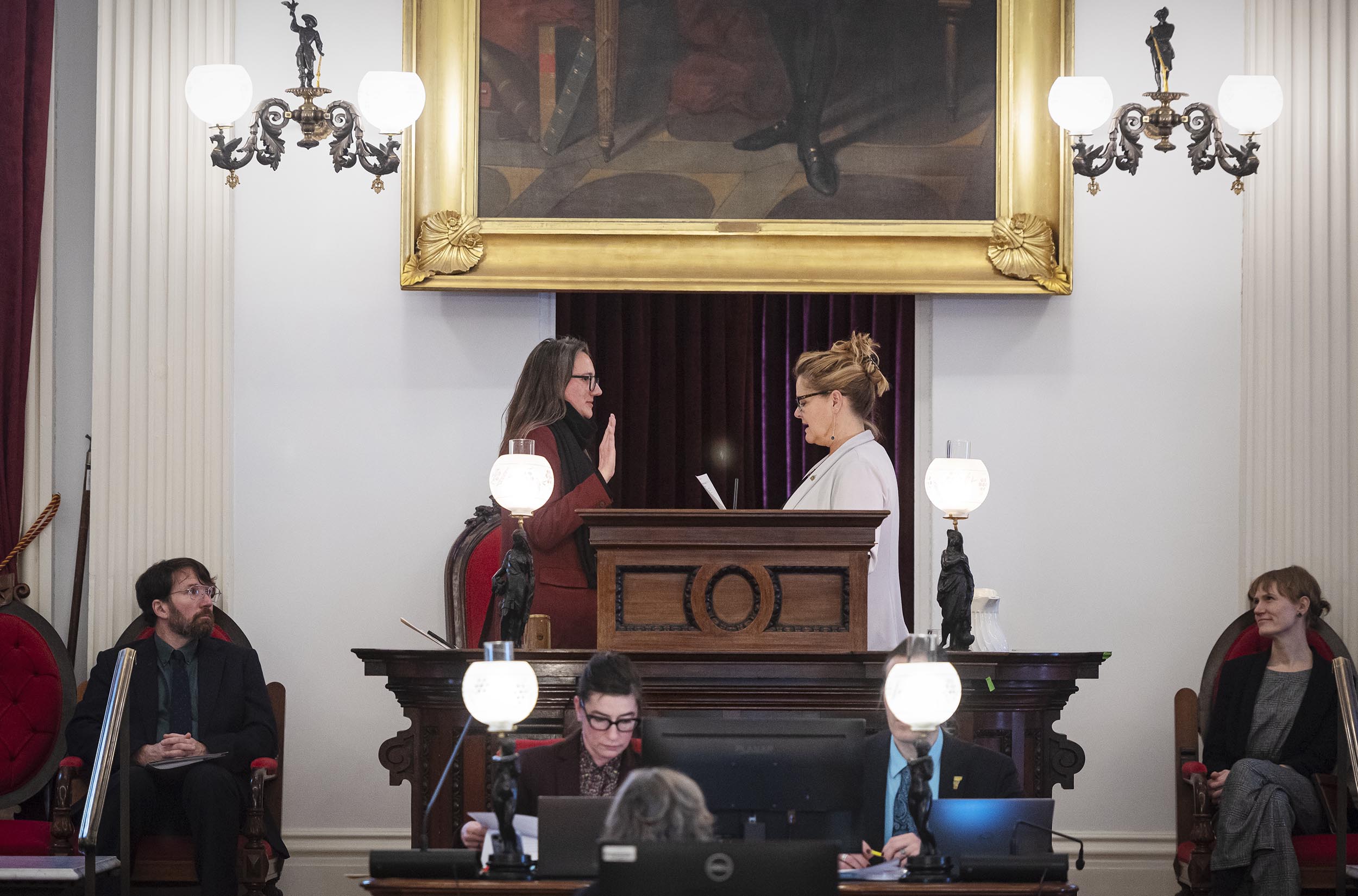 A person is being sworn in by another at a formal setting with seated individuals and a large painting in the background.