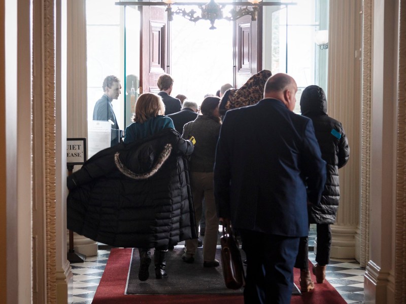 A group of people in winter attire walk towards an open door, exiting a building with tiled floors and red carpet.
