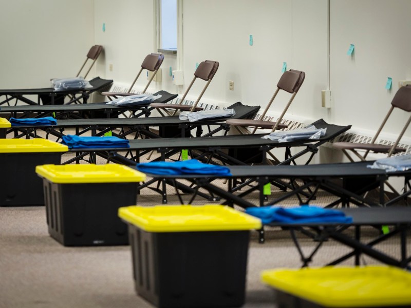 Rows of empty cots with folded blankets and yellow-lidded storage bins underneath, next to folding chairs in a plain room.