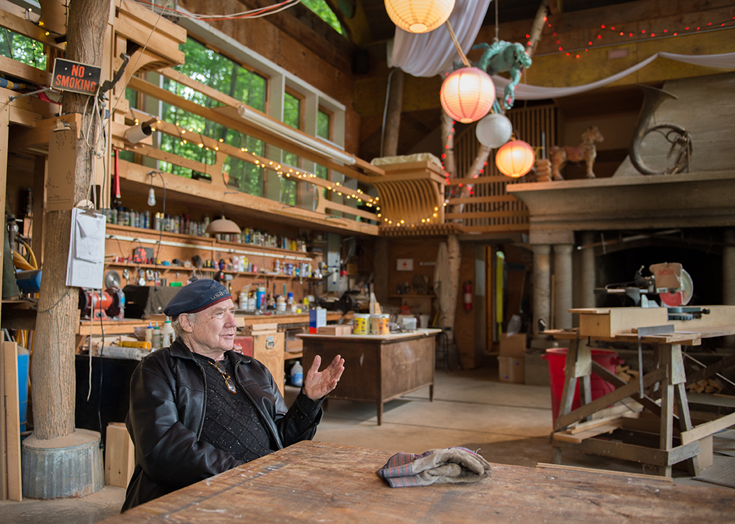 A person in a hat and jacket sits at a wooden table in a cluttered workshop with shelves, tools, and hanging lights.