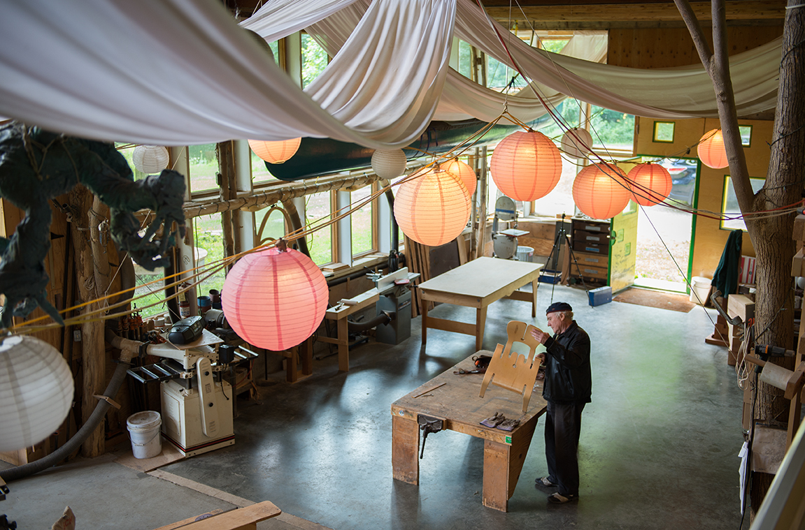A person stands in a spacious woodworking studio with hanging lanterns, tools, and large windows.