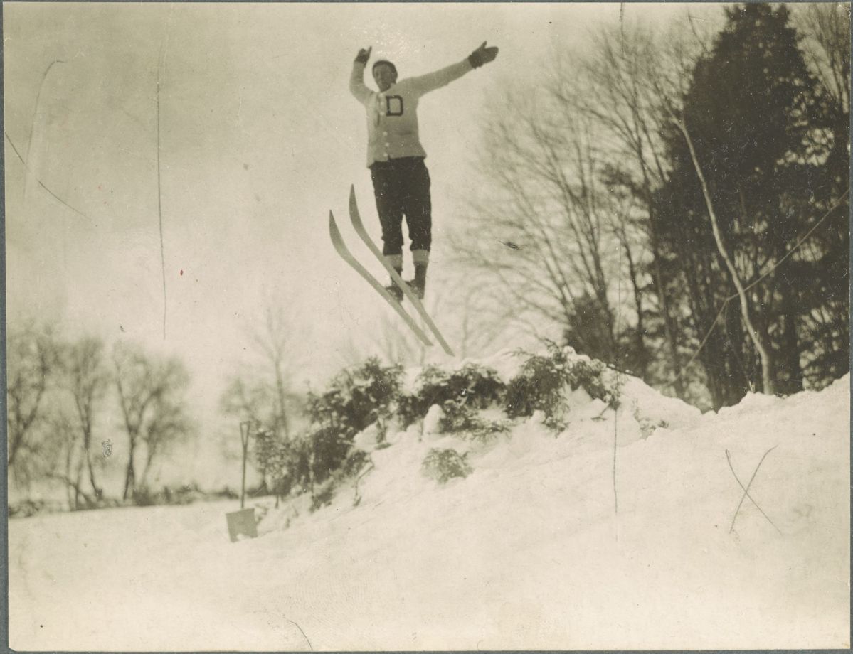 A skier mid-jump wearing a sweater with the letter "D" against a snowy backdrop with bare trees.