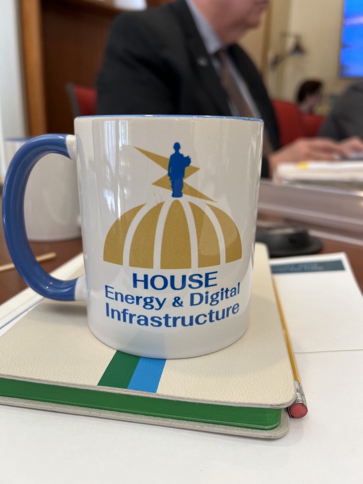 A mug displays the "House Energy & Digital Infrastructure" logo on a table. A man in the background is partially visible.