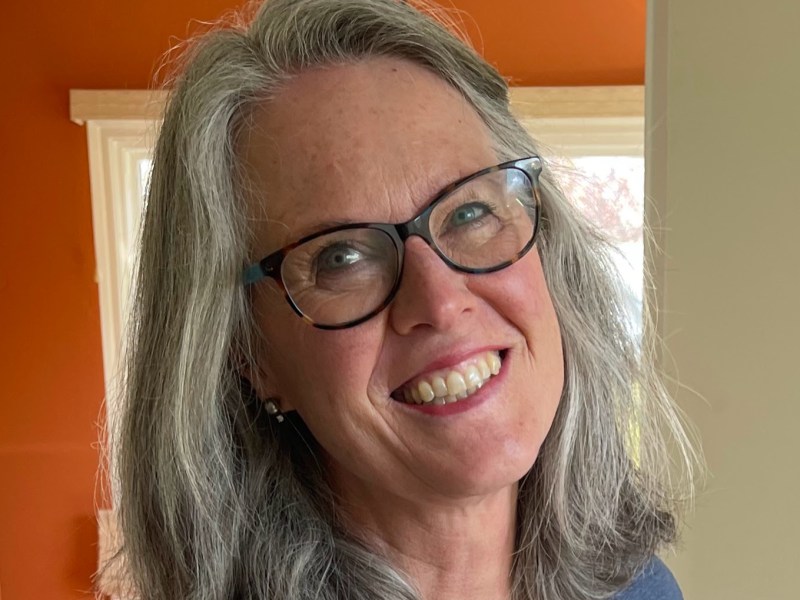 Smiling woman with glasses and gray hair stands indoors wearing a dark shirt. An orange wall and window are in the background.