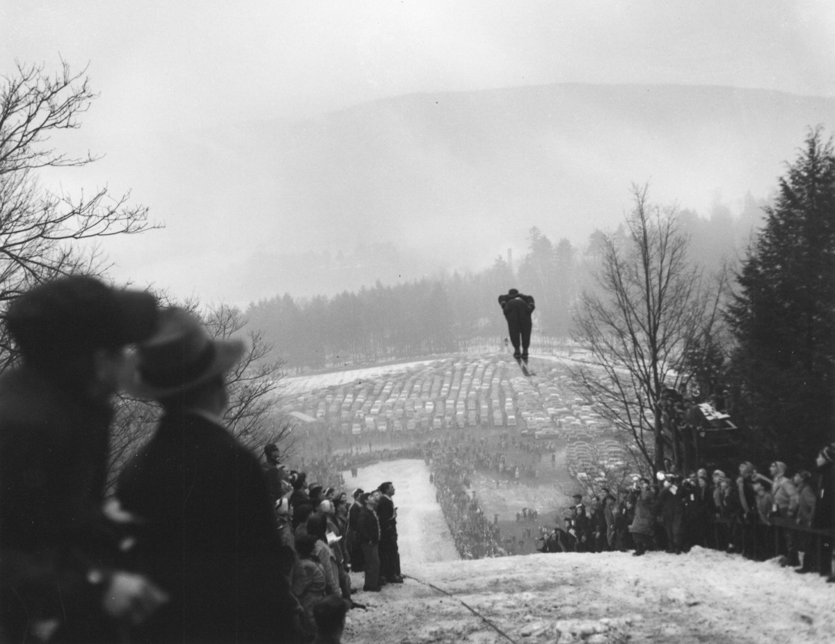 A ski jumper mid-air over a snowy slope, with spectators on both sides and a distant view of cars parked on a foggy day.