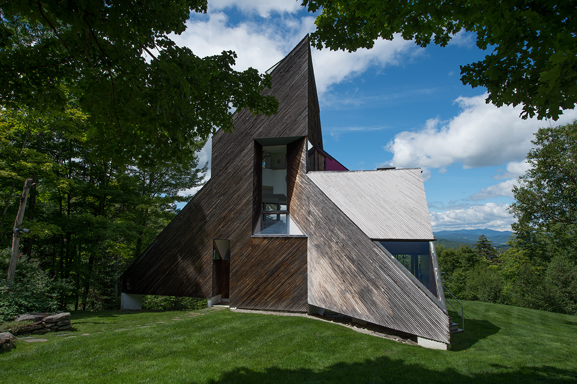 A modern triangular wooden house with large windows, surrounded by trees and grass, under a blue sky with clouds.