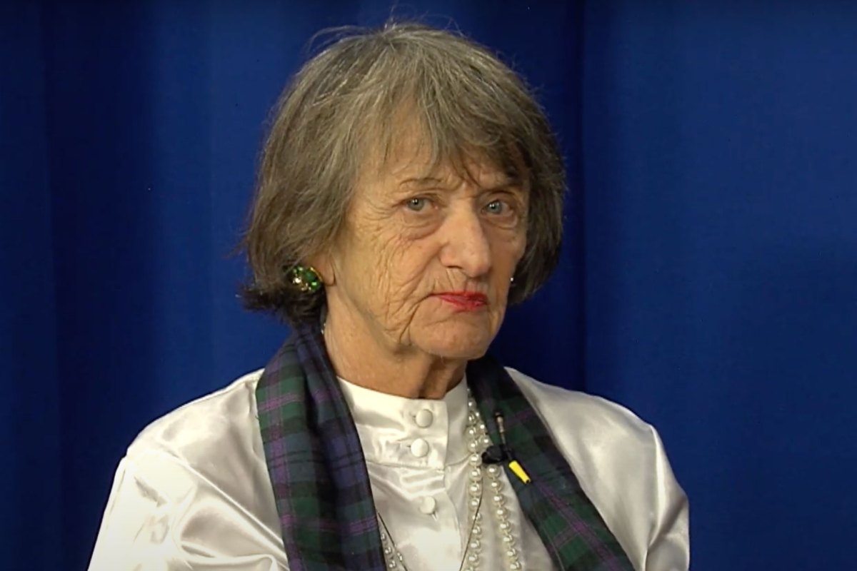 A woman with gray hair sits against a blue backdrop, wearing a white blouse, a dark plaid scarf, and green earrings.