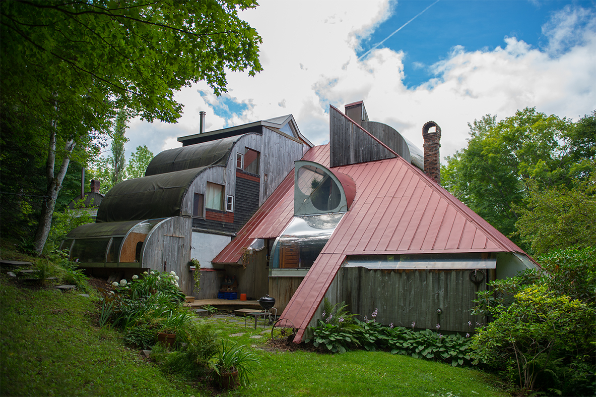 Unique multi-shaped house with geometric roofs and various textures surrounded by greenery and trees under a partly cloudy sky.