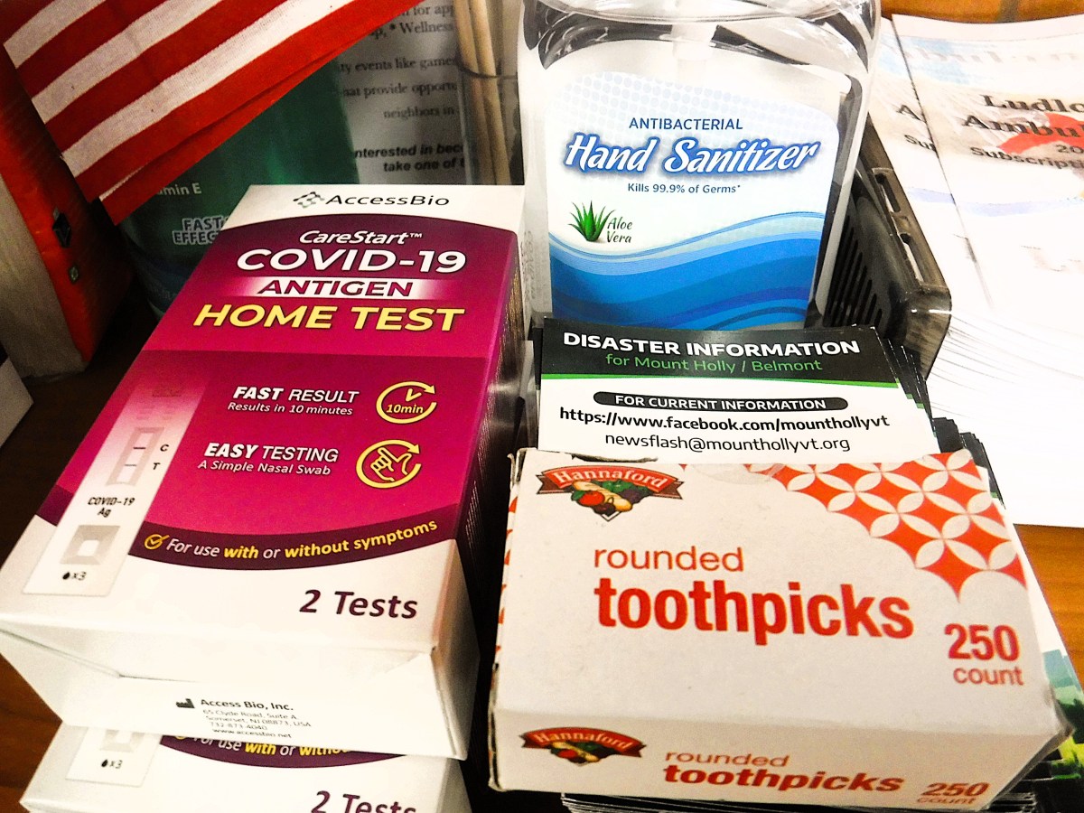 A collection of items including COVID-19 test kits, antibacterial hand sanitizer, disaster information sheet, and a box of rounded toothpicks. An American flag is partially visible in the background.