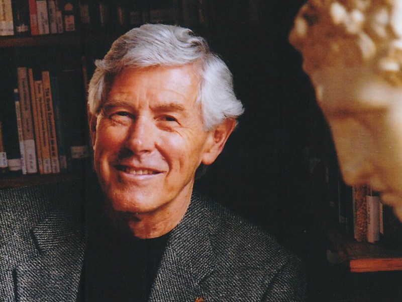A man with gray hair smiles while holding a book, surrounded by stacks of books and a bust sculpture in a library setting.