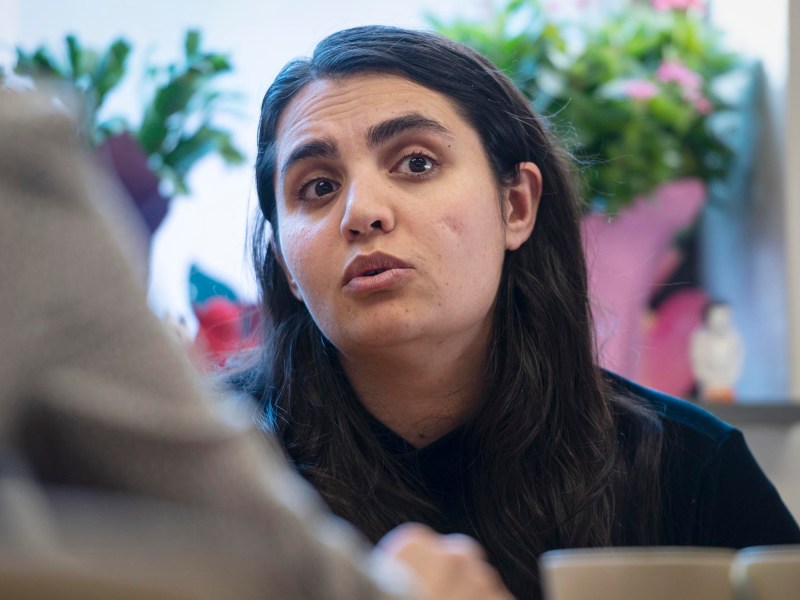 A person with long hair speaks intently indoors, with colorful flowers in the background.