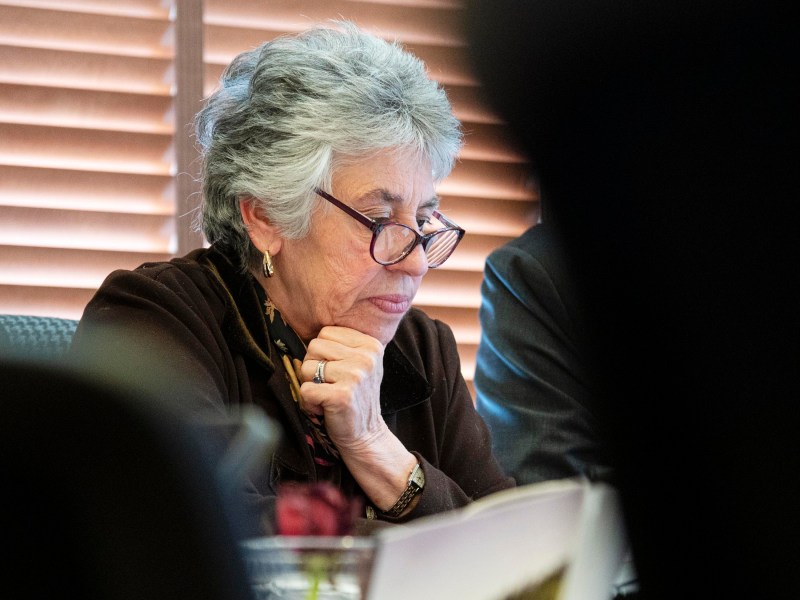 An older woman with short gray hair and glasses sits in thought, resting her chin on her hand, in front of window blinds.