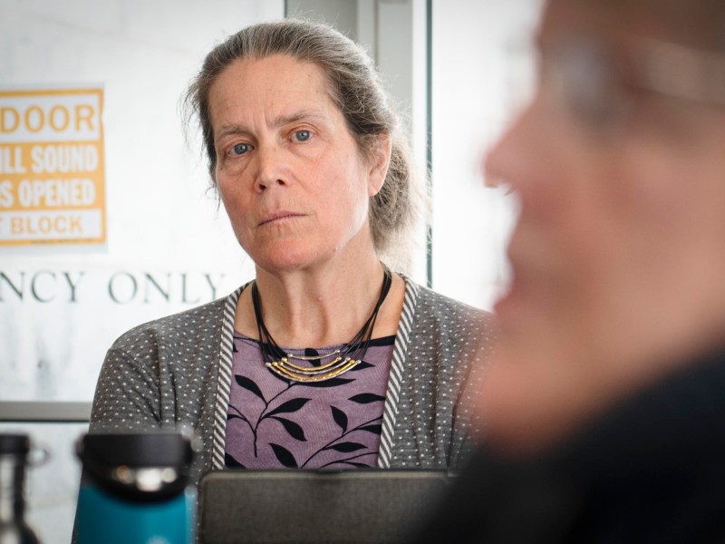 Woman with a thoughtful expression sits in a room, facing another person in the foreground. Sign on the door in the background partly visible.