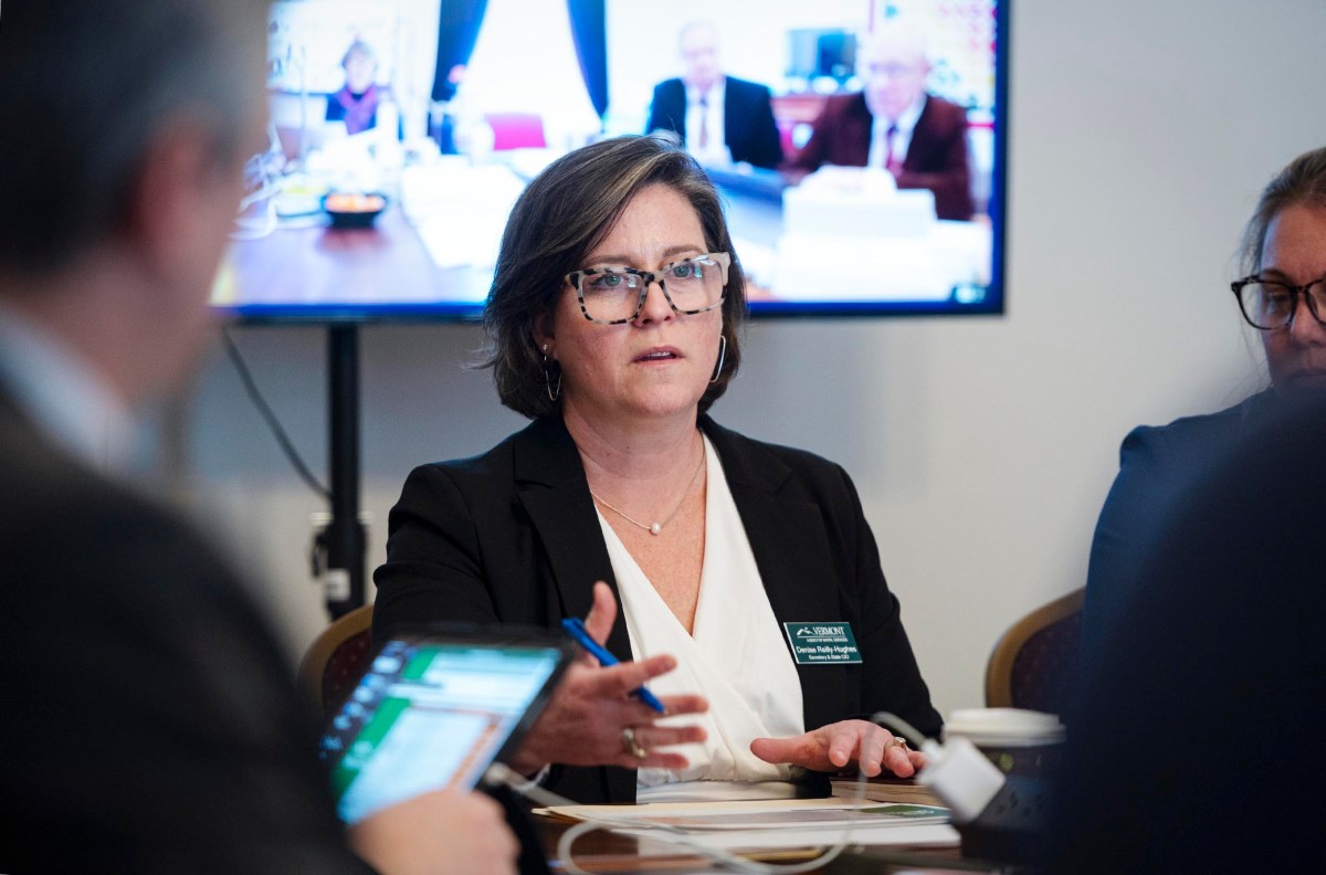 A woman in business attire speaks at a meeting with a screen in the background showing a video conference.