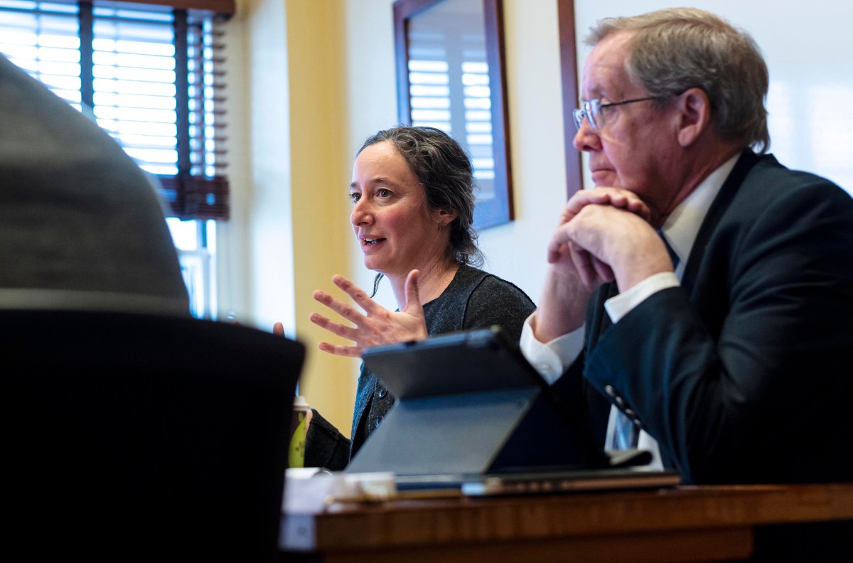 A woman and a man sit at a table in a meeting room. The woman is speaking with animated gestures while the man listens attentively with hands folded.