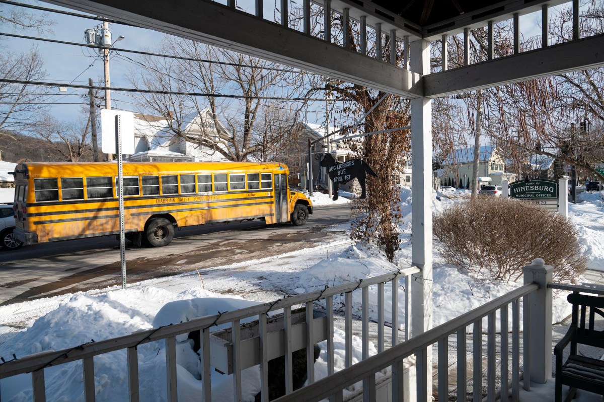 A yellow school bus drives through a snowy neighborhood intersection with houses and bare trees, viewed from a porch with white railings.