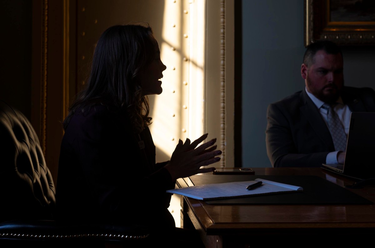 Silhouetted woman gestures while seated at a desk in an office; a man with a laptop sits nearby.