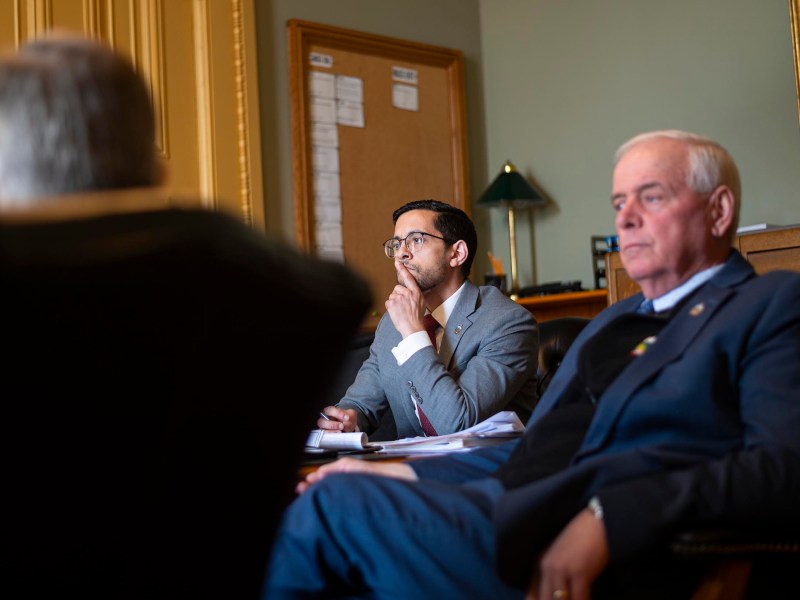 Three men in a meeting room, sitting and listening attentively. Two are in focus, wearing suits, with one holding papers and the other looking thoughtful. A notice board is in the background.