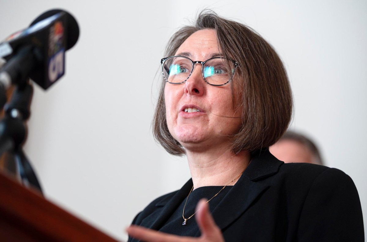 A woman in glasses speaks at a microphone, wearing a black outfit, with a blurred background.