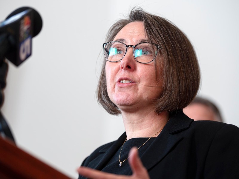 A woman in glasses speaks at a microphone, wearing a black outfit, with a blurred background.