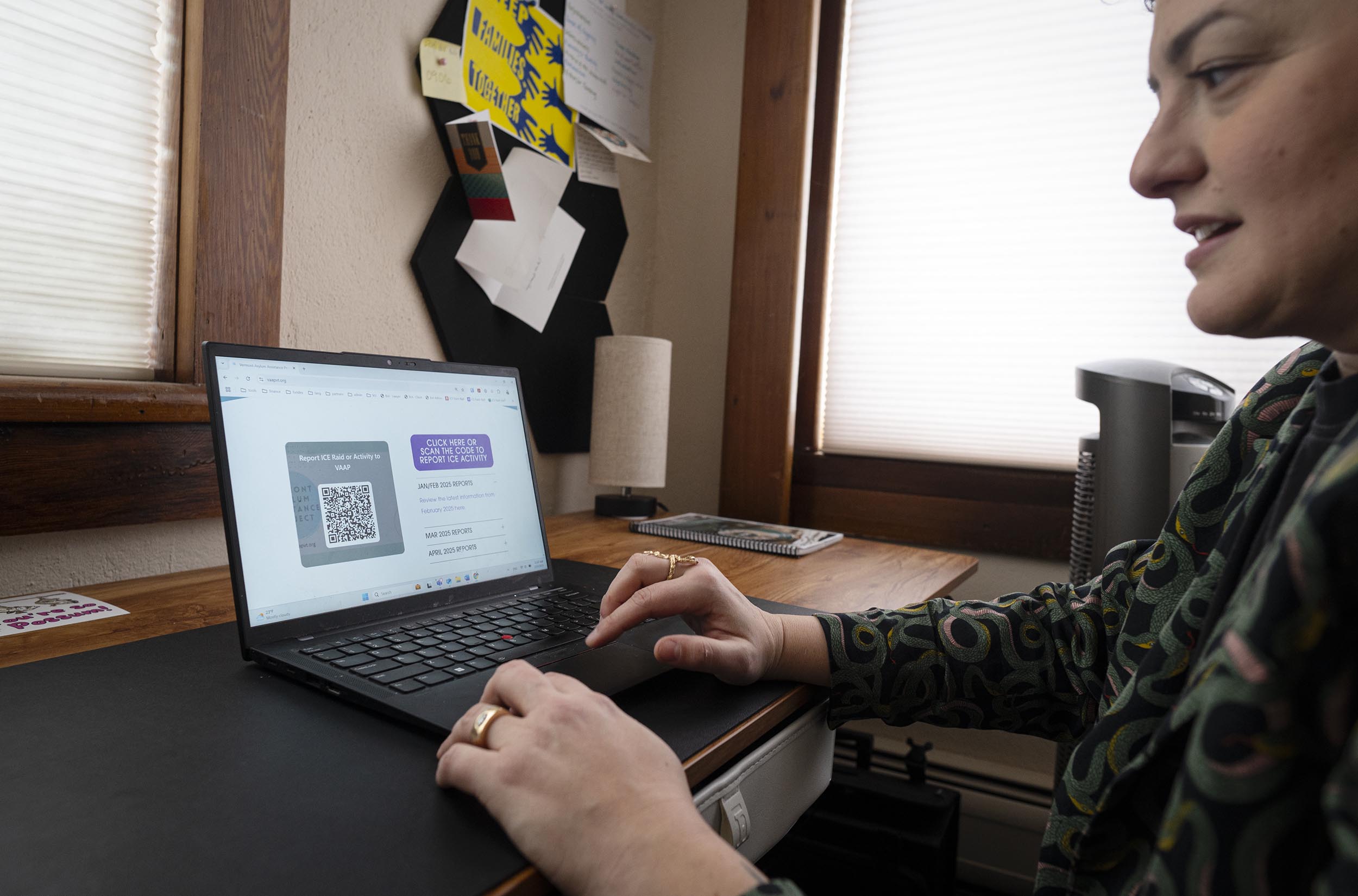 Person using a laptop, viewing a webpage displaying a QR code and text. Office environment with bulletin board and paperwork in the background.