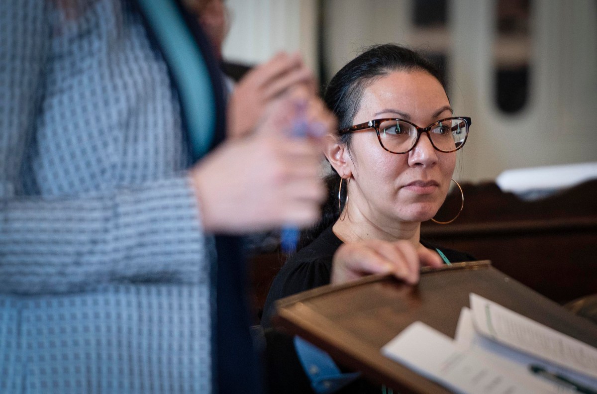 A woman wearing glasses and hoop earrings looks attentively while sitting indoors next to a standing person. Papers are visible in the foreground.