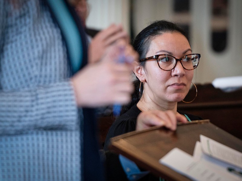 A woman wearing glasses and hoop earrings looks attentively while sitting indoors next to a standing person. Papers are visible in the foreground.