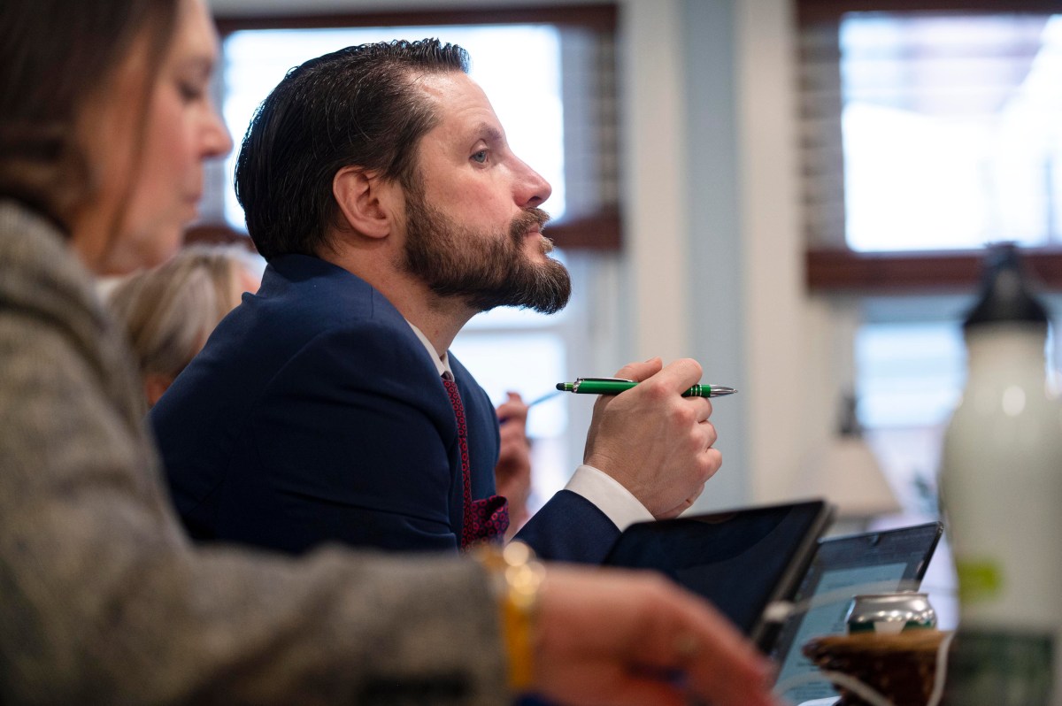 Man in a suit attentively listens in a meeting, holding a pen. Another person is partially visible in the foreground.