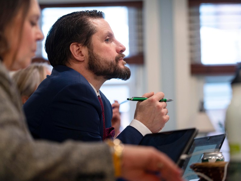 Man in a suit attentively listens in a meeting, holding a pen. Another person is partially visible in the foreground.