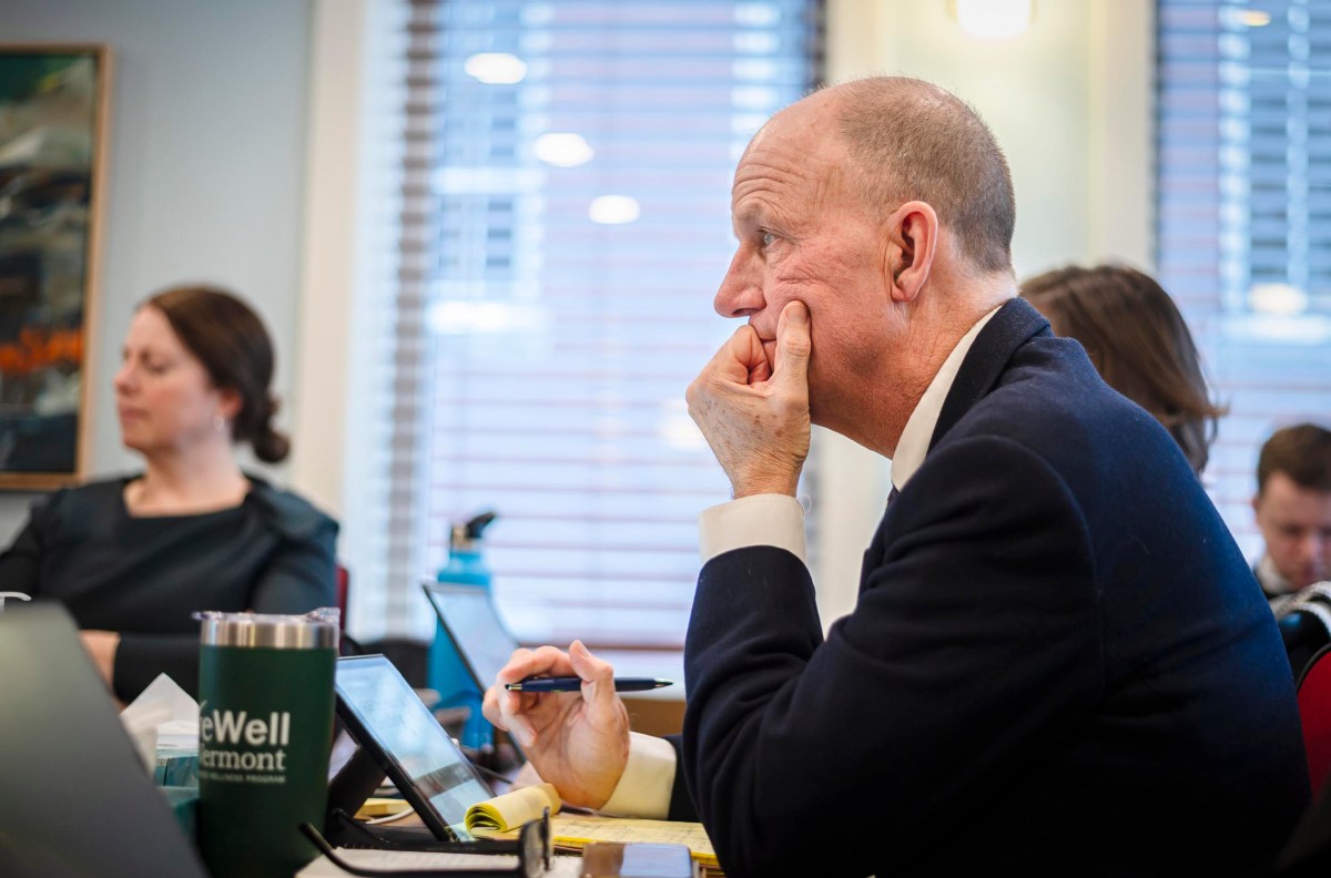 A man in a suit sits at a table, resting his chin on his hand, focused on a discussion. A woman and another person are visible in the background. Various items are on the table.