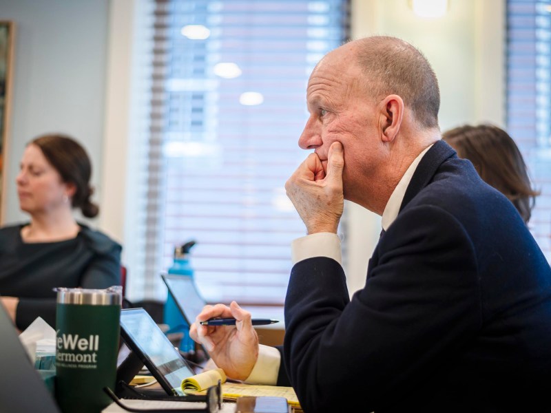 A man in a suit sits at a table, resting his chin on his hand, focused on a discussion. A woman and another person are visible in the background. Various items are on the table.