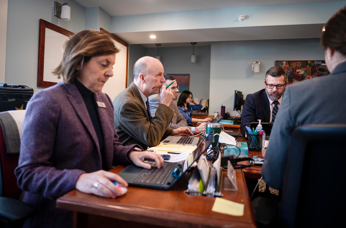 People sitting at a conference table, focused on documents and laptops. Office setting with papers and pens visible.