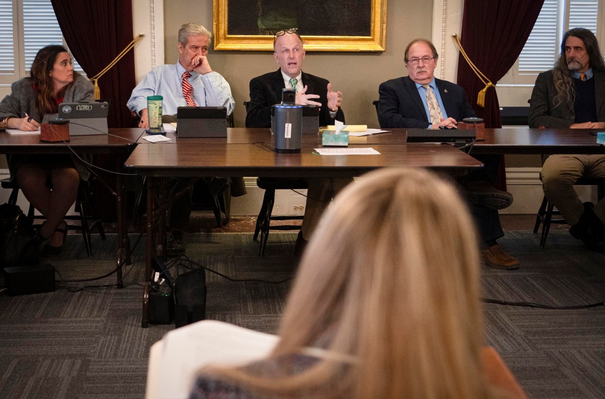 A group of five people sitting at a conference table, with one person speaking and gesturing. Another person is sitting opposite them, seen from the back.
