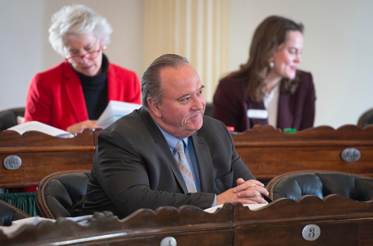 A man in a suit sits at a desk, listening, with two women seated behind him. Papers are on the desk.