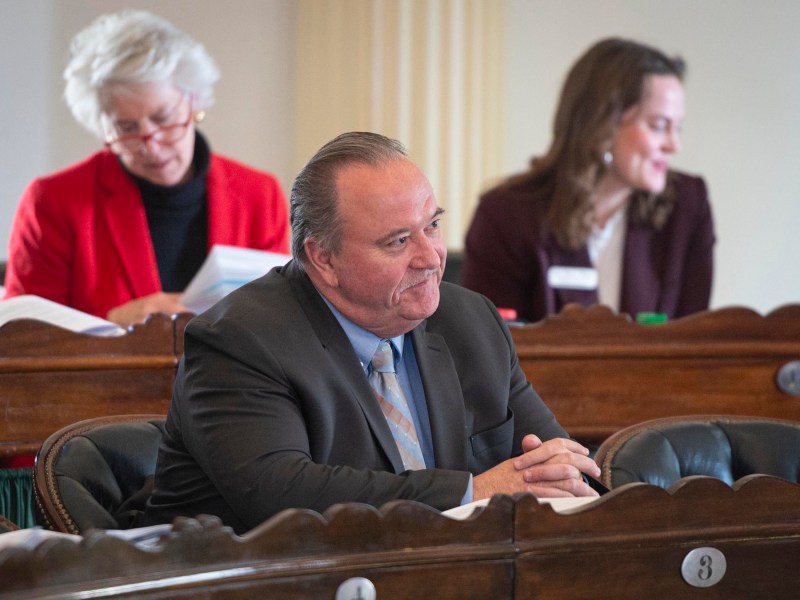 A man in a suit sits at a desk, listening, with two women seated behind him. Papers are on the desk.