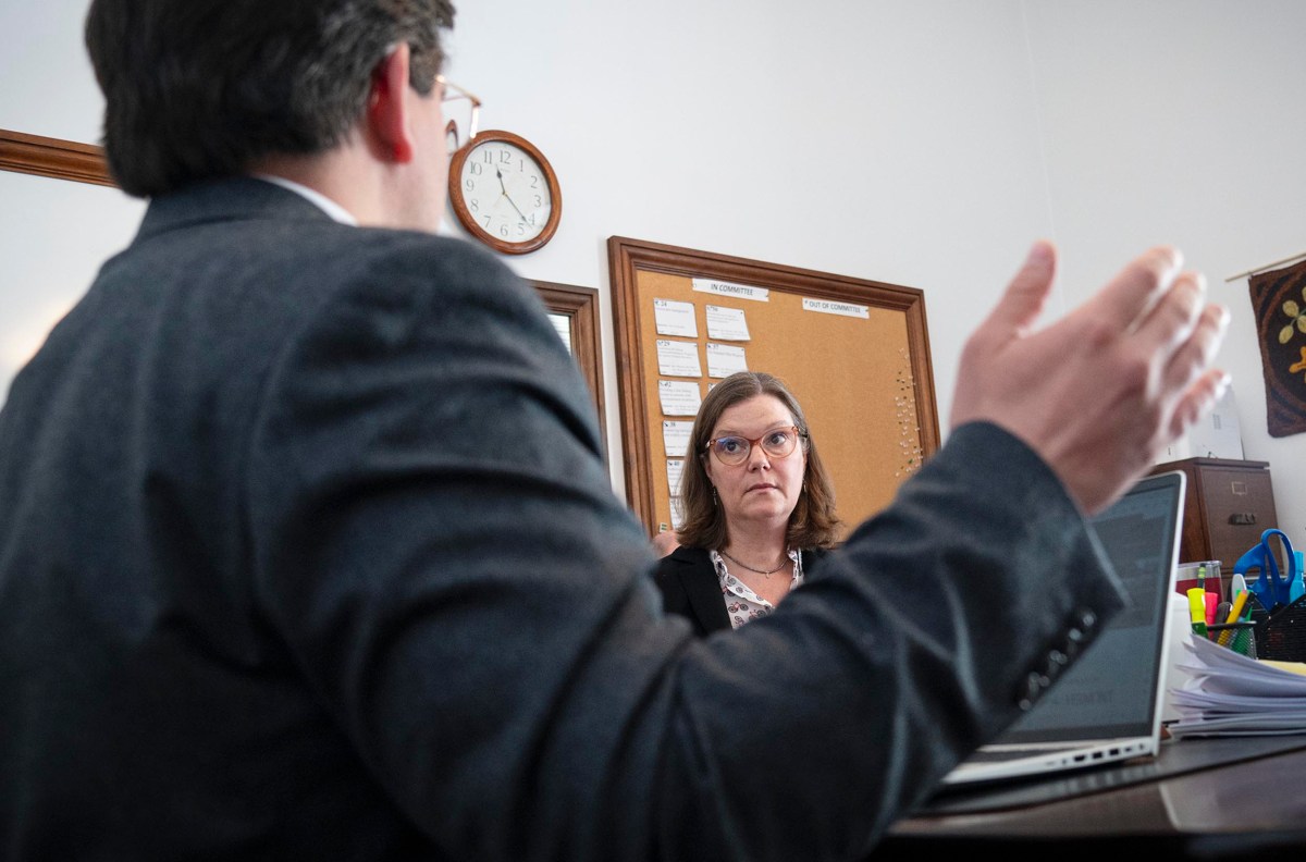 Two people in a meeting. A man gestures in the foreground, facing a woman who listens attentively. A clock and bulletin board are visible on the wall.