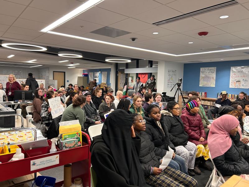 A group of people seated in a room attentively listening, with some holding papers. A cart with snacks is visible in the foreground.