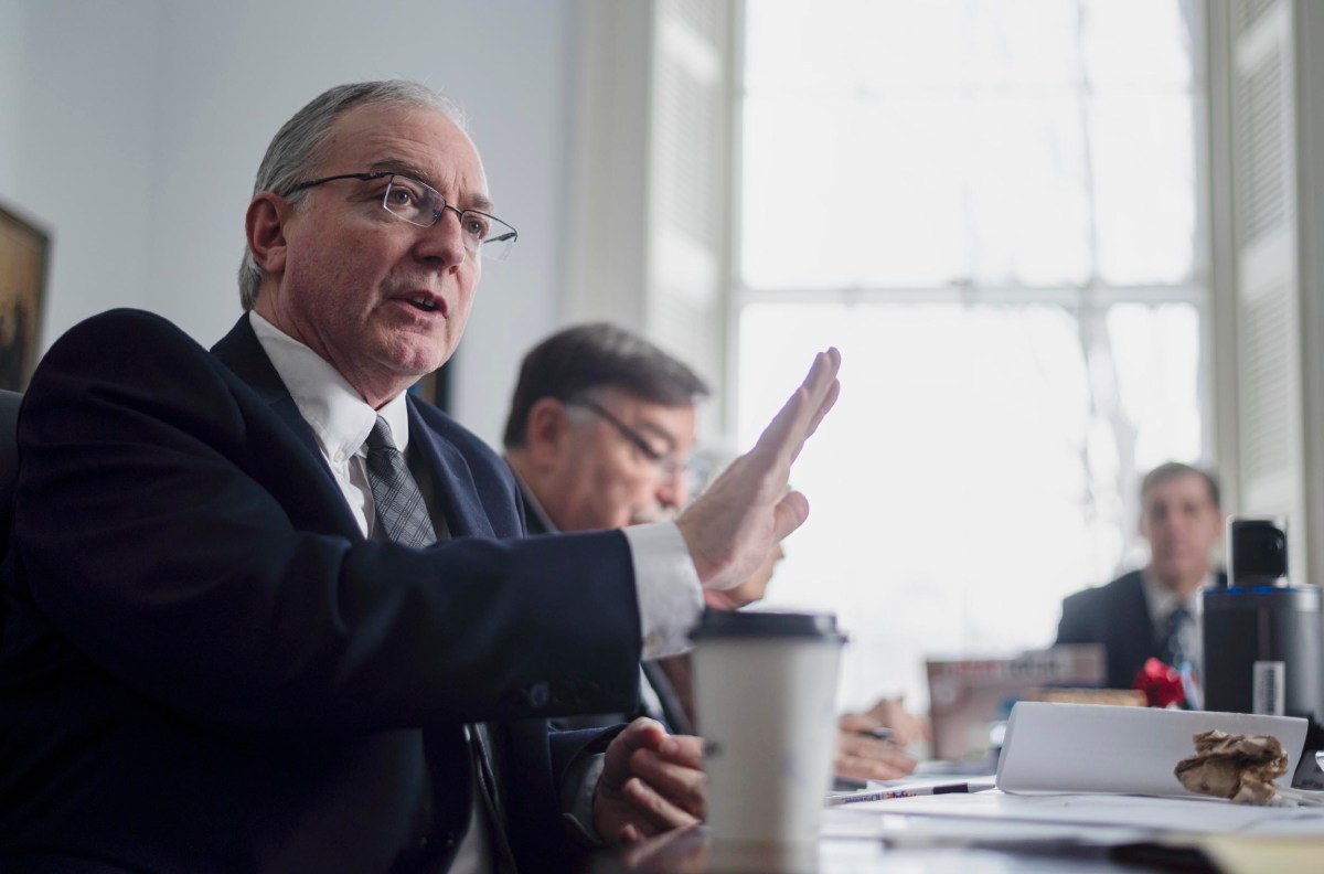A man in a suit gestures while speaking at a meeting table. Two other men are seated nearby, with papers and coffee cups on the table. A large window is in the background.