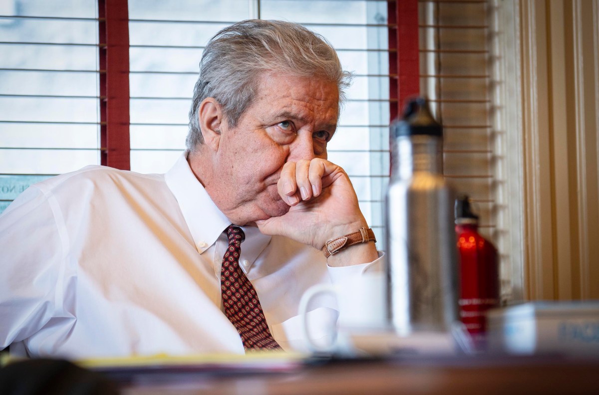 Man in a white shirt and patterned tie sits at a table, resting his chin on his hand, looking pensive. A bottle and documents are visible in the foreground.