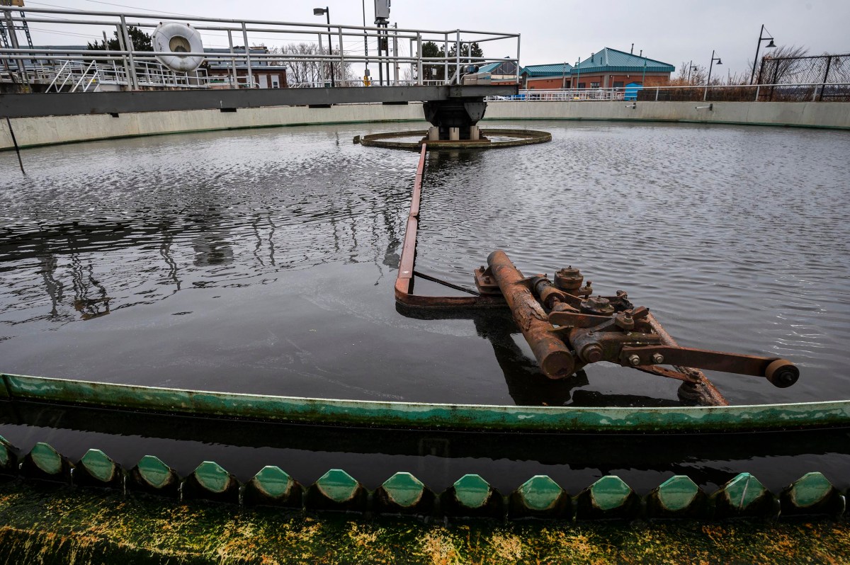 A circular wastewater treatment basin with a mechanical arm extends across the surface. Buildings and a life preserver are visible in the background.