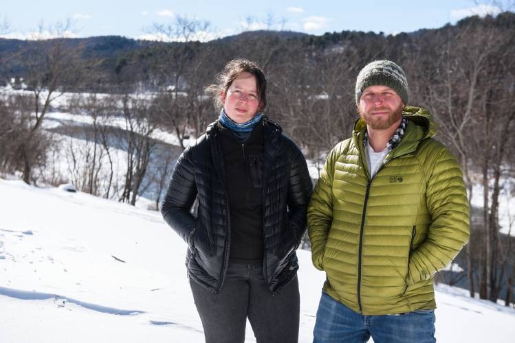 Two people stand in a snowy landscape, both wearing jackets. The background features snow-covered ground and bare trees under a clear sky.