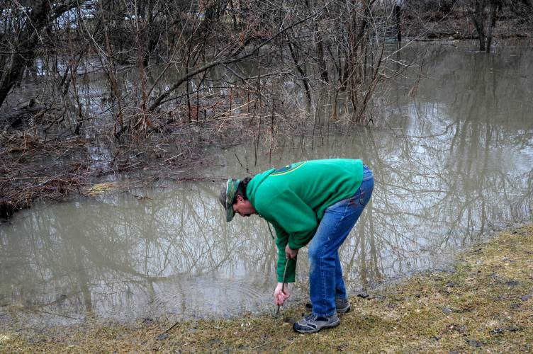 A person bends down near flooded water, dressed in a green hoodie and jeans, with leafless trees in the background.