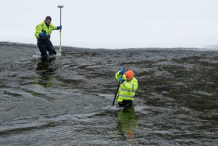 Two people in high-visibility jackets stand in a shallow, flowing river, taking measurements with poles. Snow is visible on the banks.