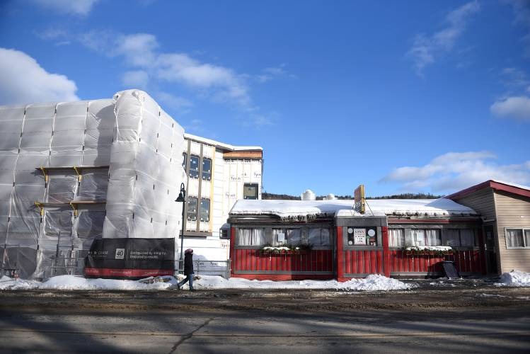 A red diner with a sign reading "Noon Mark Diner" is situated on a snowy street. To the left, a building is covered with protective wrap. A person walks on the sidewalk.