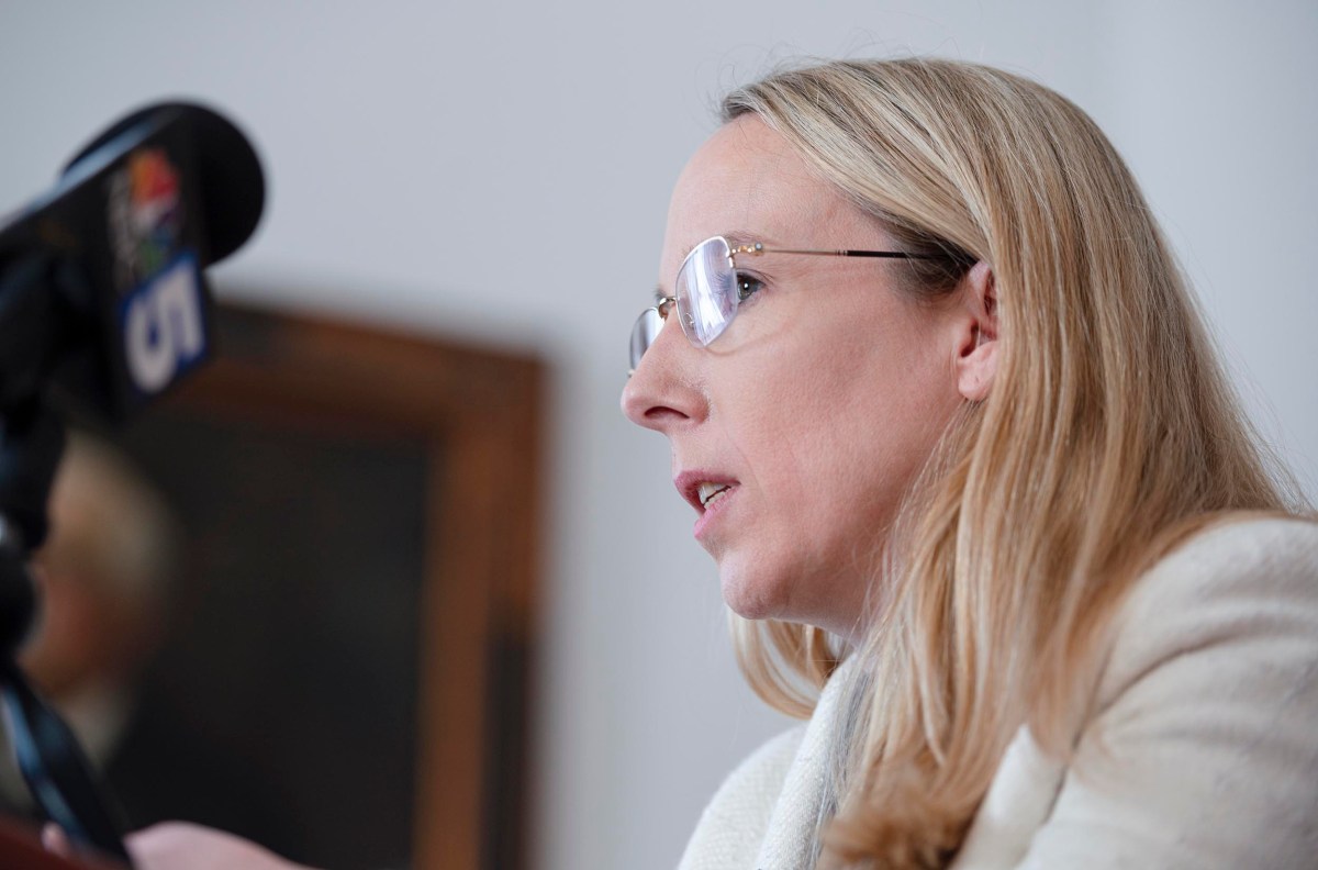 A woman with long blonde hair and glasses speaks into microphones at a podium.